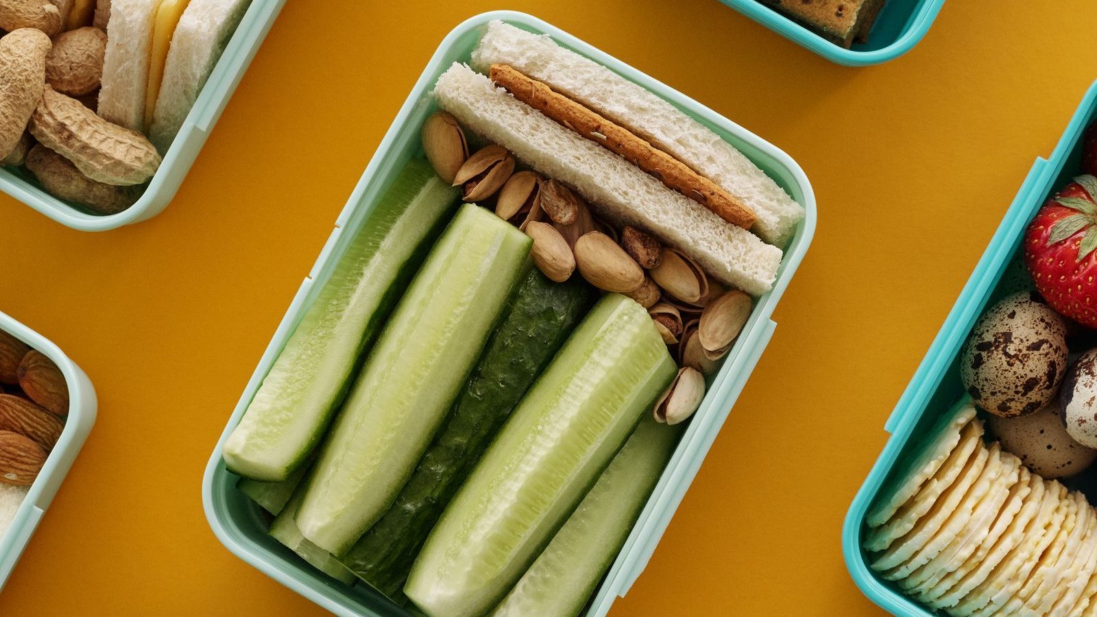 An overhead view of several lunch boxes packed with sandwiches, cheese, fruit and cucumber