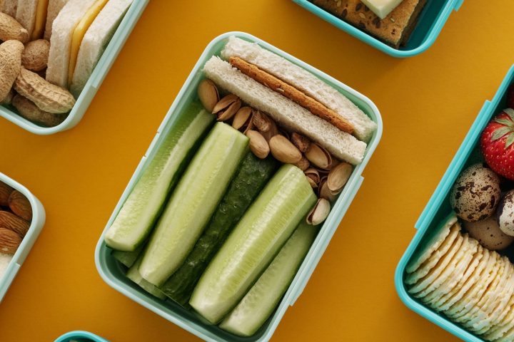 An overhead view of several lunch boxes packed with sandwiches, cheese, fruit and cucumber