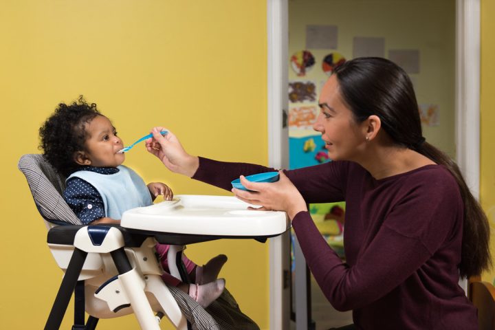 An adult spoon-feeding a smiling baby who is wearing a bib and sitting in a high chair
