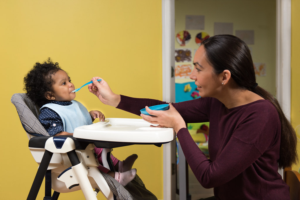 An adult spoon-feeding a smiling baby who is wearing a bib and sitting in a high chair