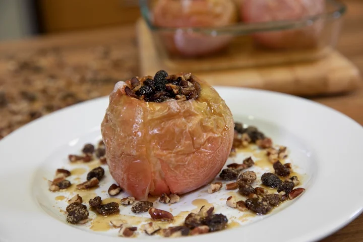 A soft baked apple stuffed with raisins, surrounded by pecans and syrup on a white plate
