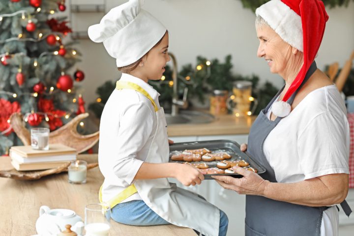A child in a chef's hat and a grandmother holding a tray of gingerbread cookies beside a Christmas tree