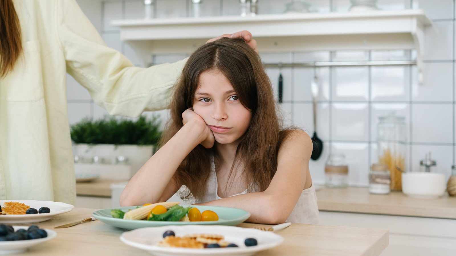 A young girl resting her chin on her hand, looking unenthusiastically at a plate of food