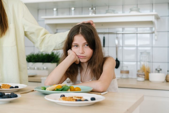 A young girl resting her chin on her hand, looking unenthusiastically at a plate of food