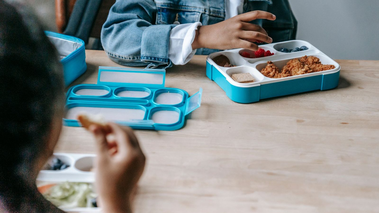 Two children sitting at a table eating from compartment lunch boxes