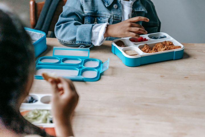 Two children sitting at a table eating from compartment lunch boxes
