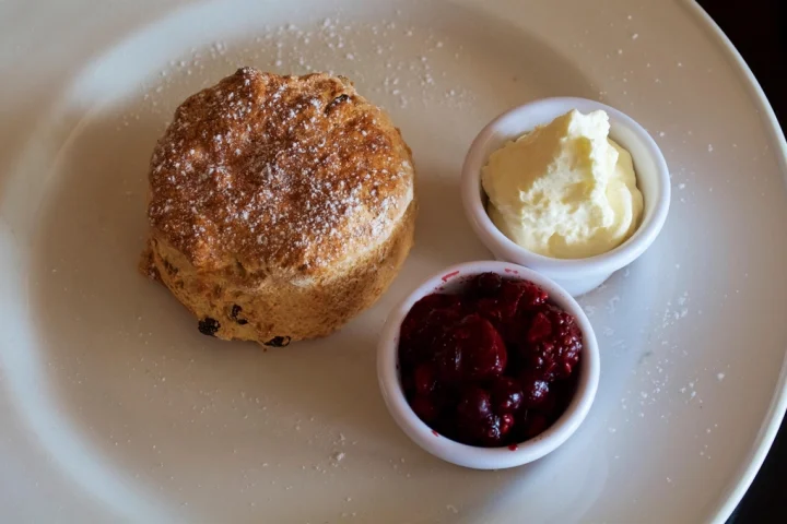 A scone on a plate beside small pots of clotted cream and raspberry jam