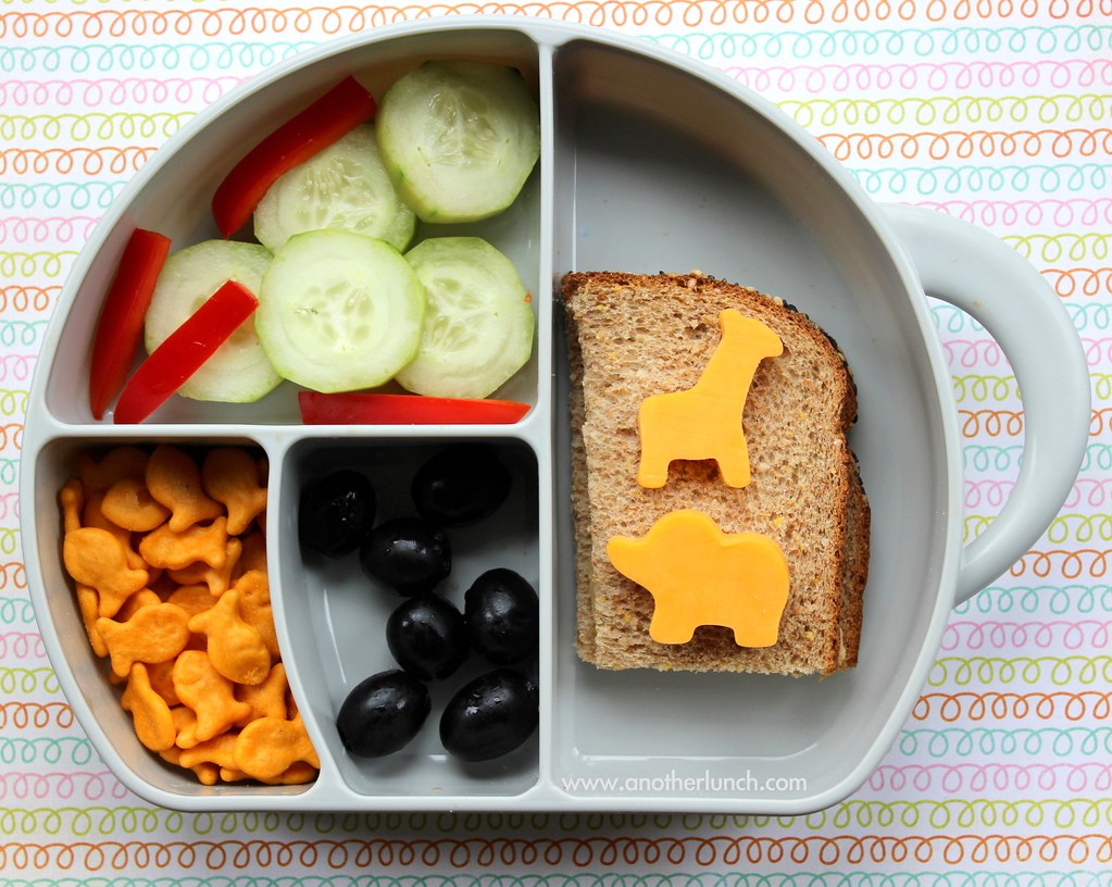 A grey divided children's plate with cucumber, red pepper, crackers, olives and bread topped with animal-shaped cheese