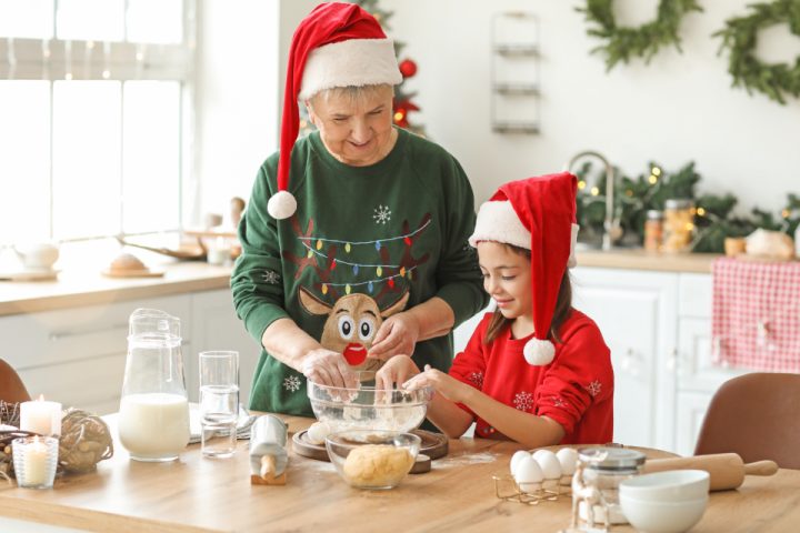 A grandmother and granddaughter in Santa hats mixing ingredients in a glass bowl in a festive kitchen