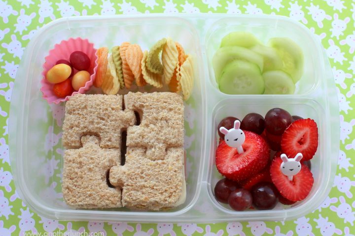 A child's bento-style lunchbox with a puzzle-shaped sandwich, cucumber, strawberries, grapes and crackers