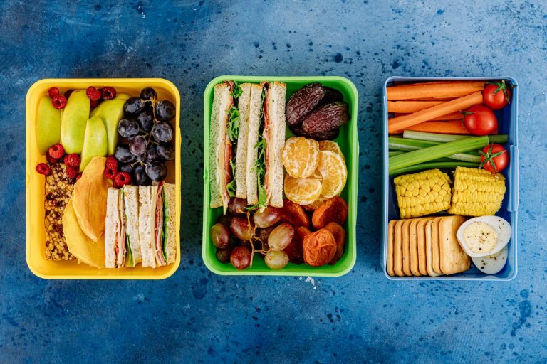 Three packed bento lunch boxes holding sandwiches, fruit and raw vegetables