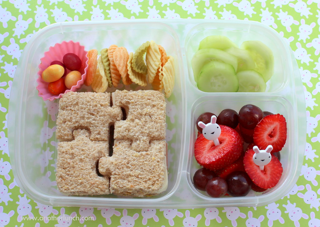 A child's bento-style lunchbox with a puzzle-shaped sandwich, cucumber, strawberries, grapes and crackers
