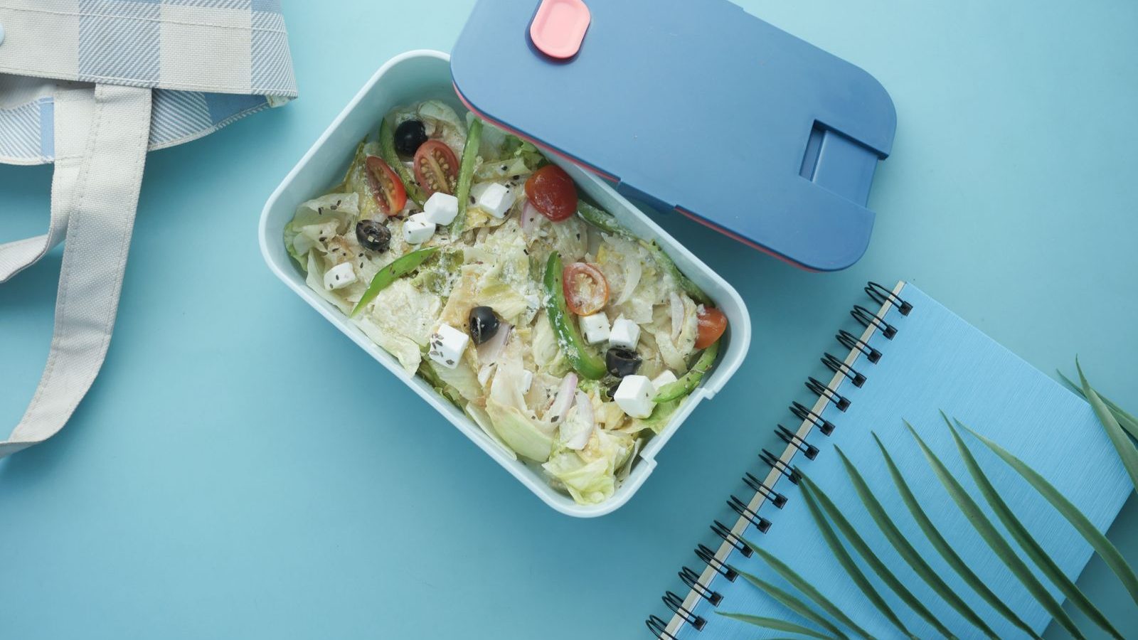 A packed lunch box of salad beside a cool bag and notebook on a blue surface