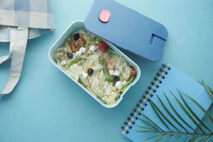 A packed lunch box of salad beside a cool bag and notebook on a blue surface