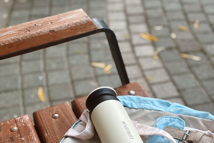 A stainless steel reusable water bottle resting on a backpack on a wooden bench