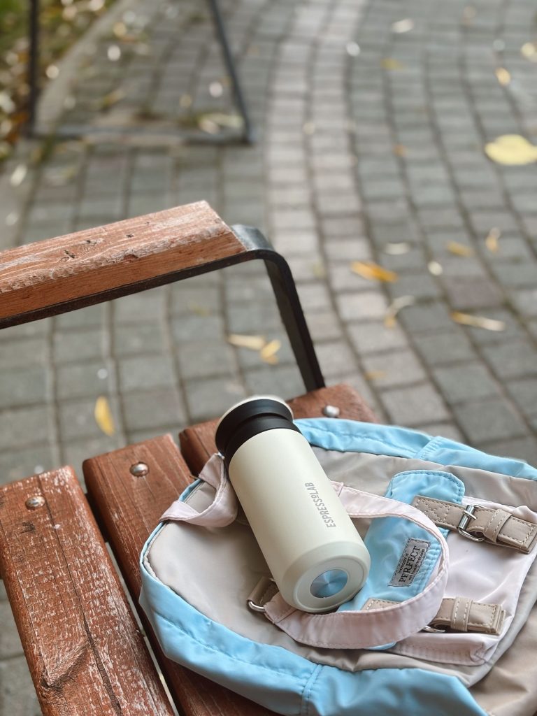 A stainless steel reusable water bottle resting on a backpack on a wooden bench