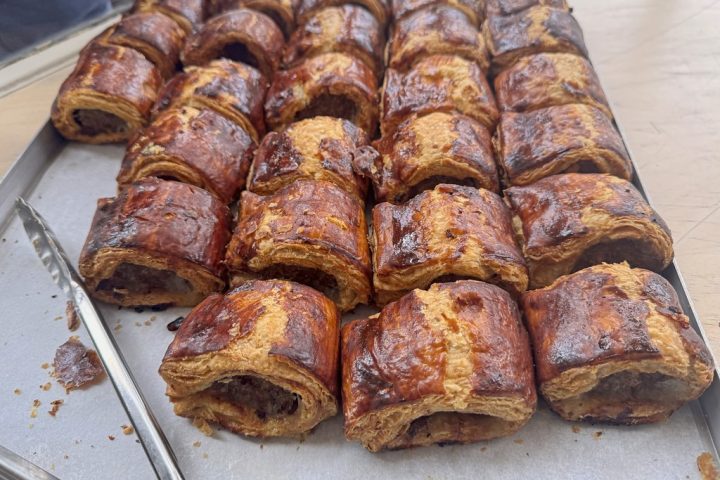 A tray of golden, flaky puff pastry sausage rolls with serving tongs