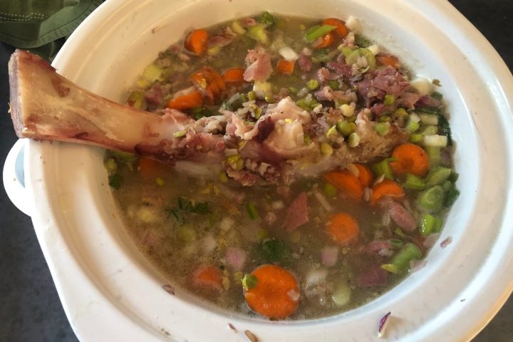 A white slow cooker filled with split pea soup, carrots and a ham bone