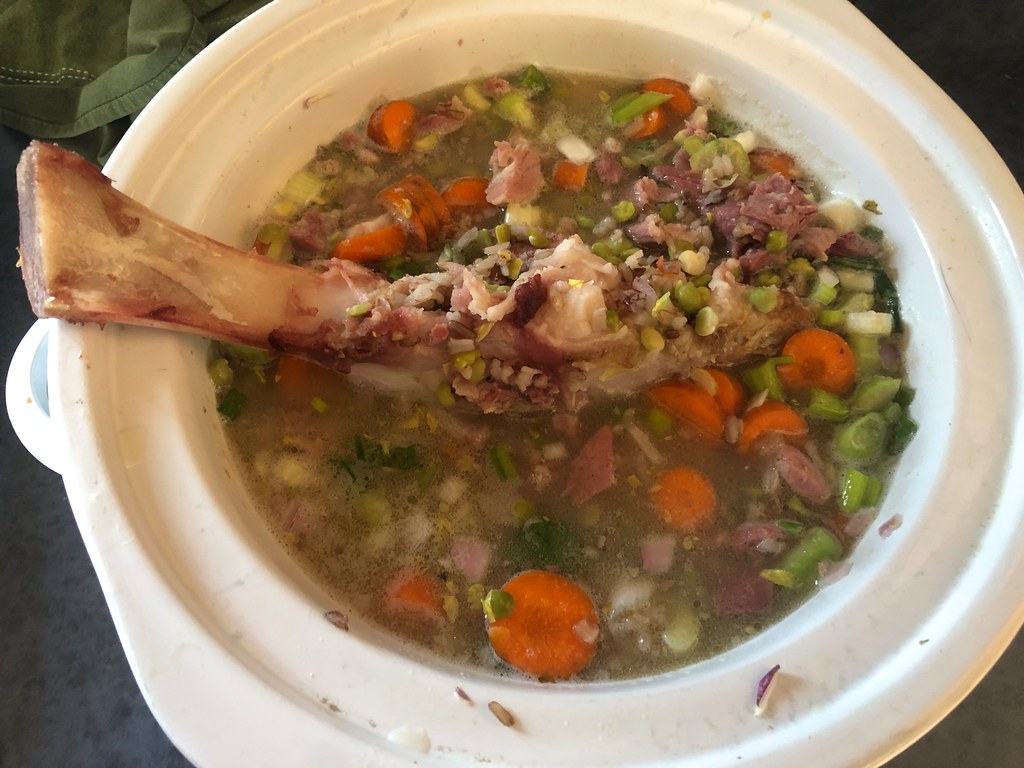 A white slow cooker filled with split pea soup, carrots and a ham bone