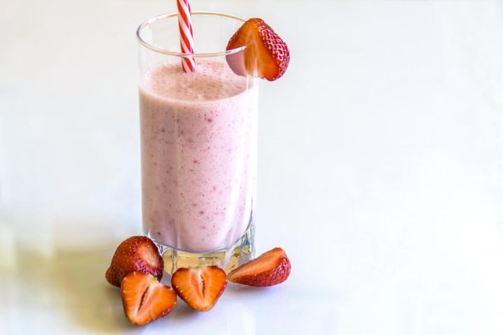 A pink strawberry smoothie in a tall glass with a striped straw, beside fresh strawberries