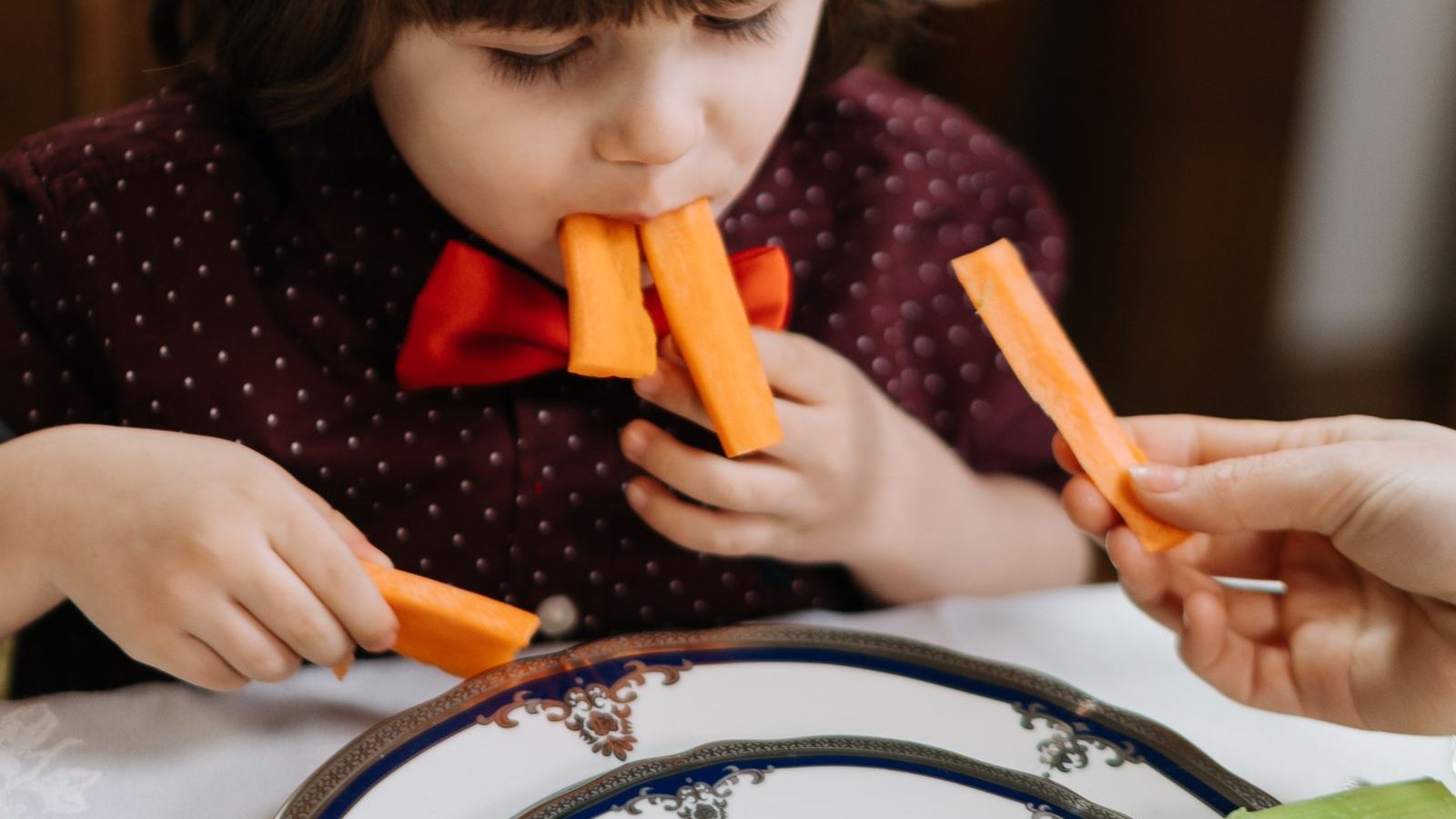 A small child eating carrot sticks from a plate at a dining table