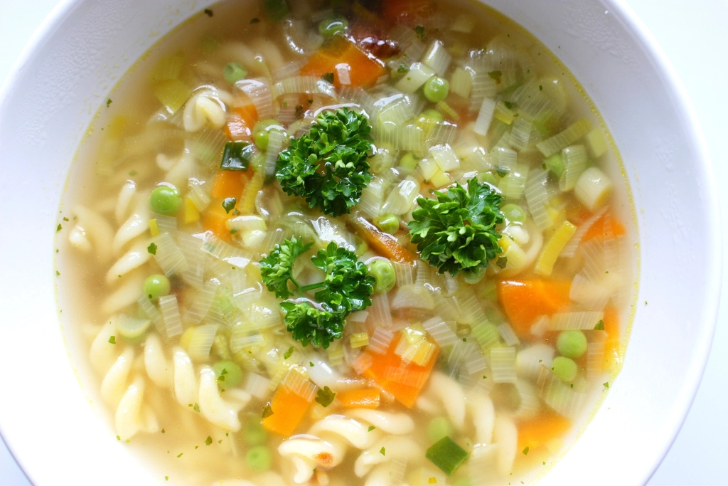 A bowl of clear vegetable soup with pasta, carrot, peas and broccoli
